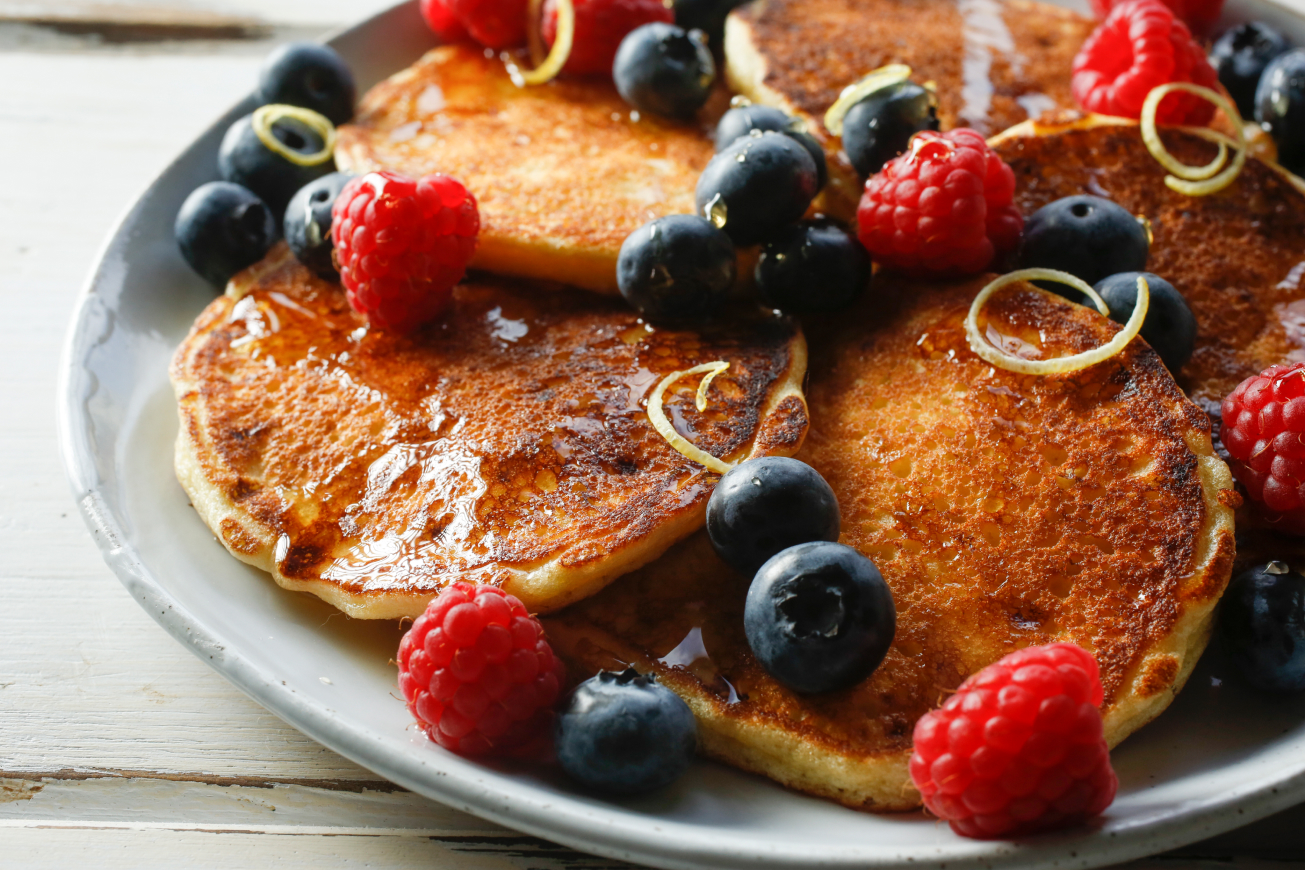 Five small pancakes on a plate topped with raspberries, blueberries, lemon zest and syrup.