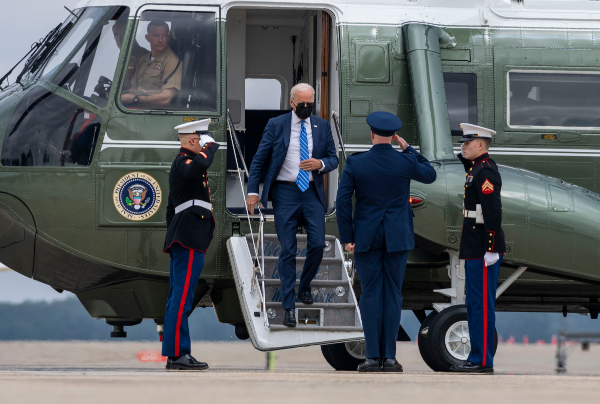 President Joe Biden, wearing a medical mask, walking down a flight of stairs out of a green helicopter. Three men in uniform salute him with their right arms from the ground.