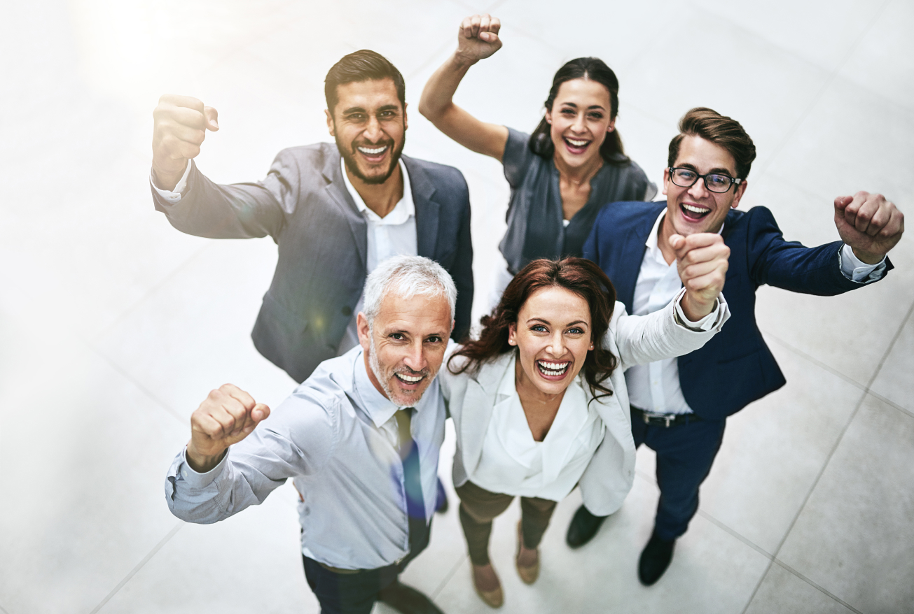 A diverse group of five people in office attire looking up at the camera, smiling, raising their fists.