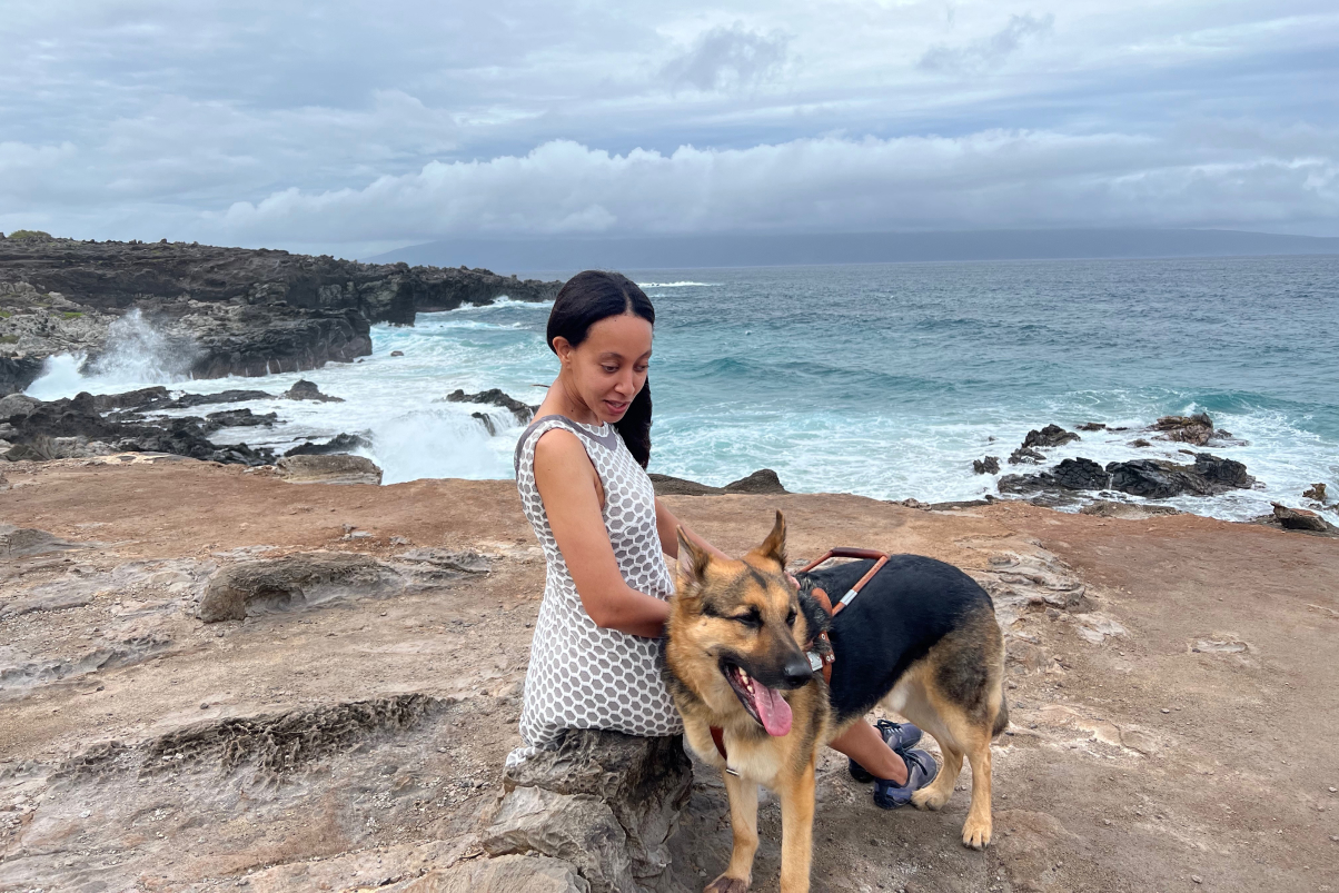 A person and a dog sitting on a cliff with the ocean behind.