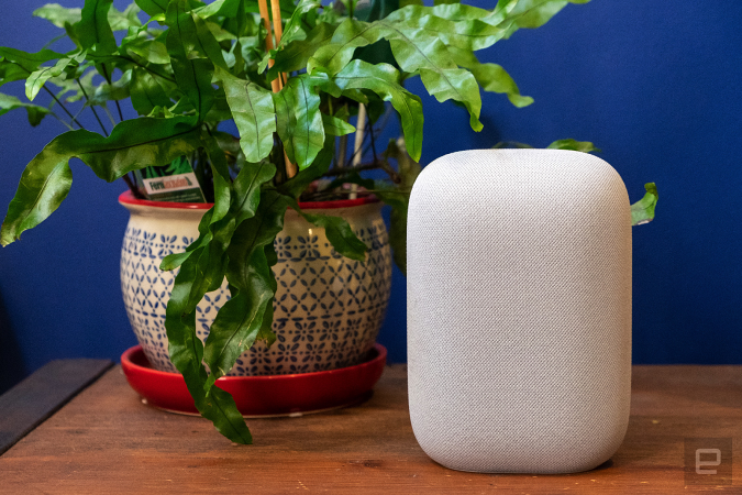 Google Nest Audio smart speaker sitting on a wooden tablet next to a green plant, against a blue wall.