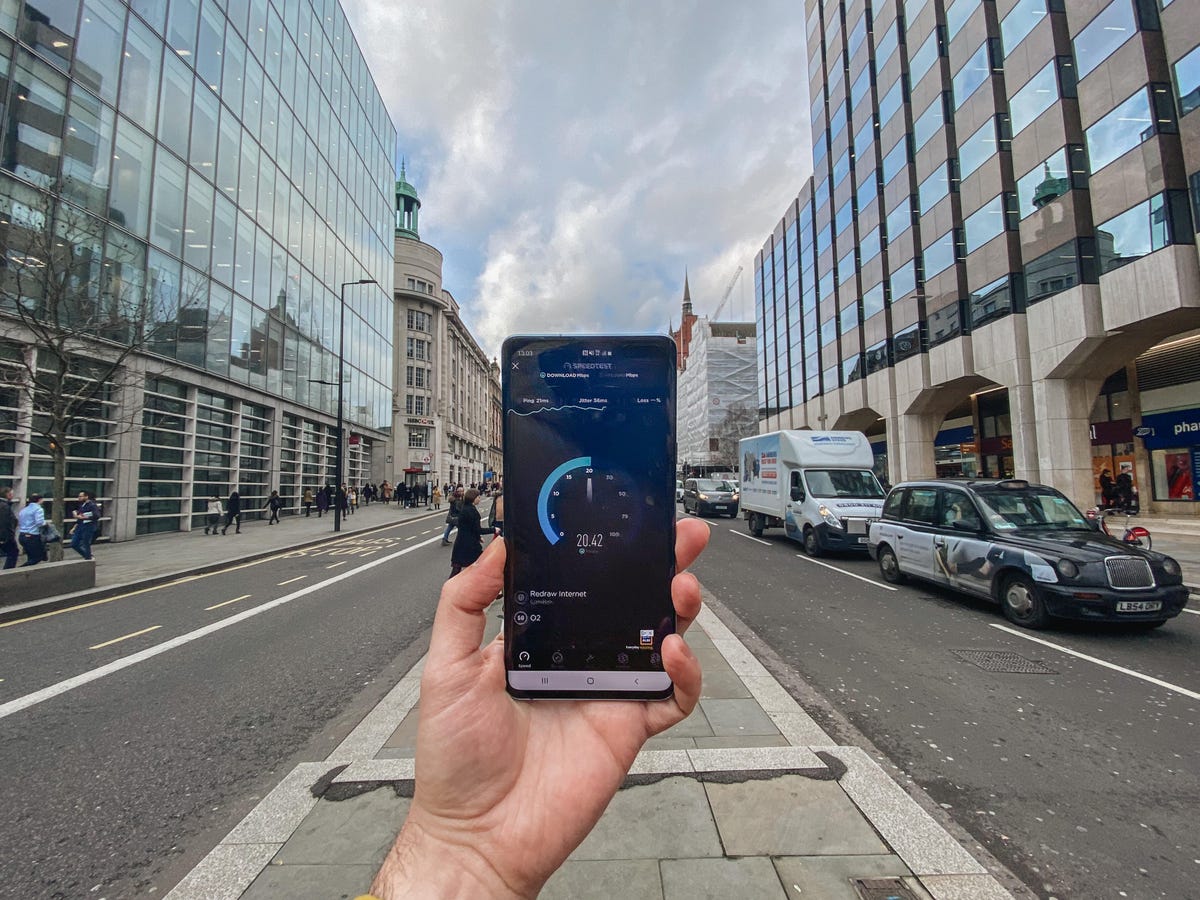 Hand holding a smartphone with street and glass buildings in background.