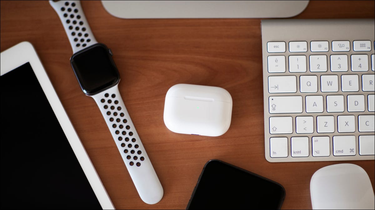 An AirPods Pro case on a wood tabletop surrounded by other Apple devices.