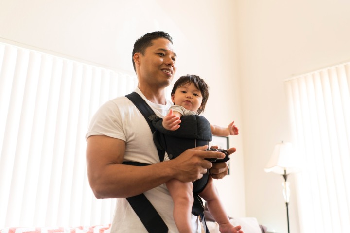 A dad standing and gaming with a baby in a carrier.